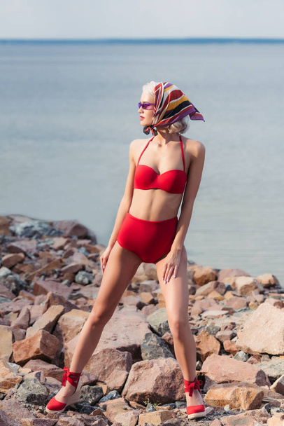 attractive elegant girl posing in red bikini and silk scarf on rocky beach at sea - Fotó, kép