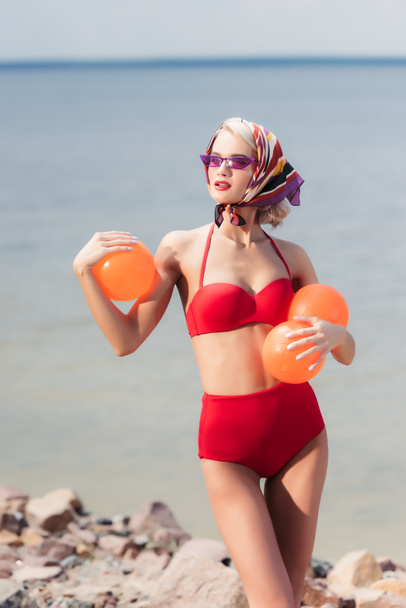mulher atraente em biquíni vermelho vintage, óculos de sol e cachecol de seda posando com bolas laranja perto do mar
 - Foto, Imagem