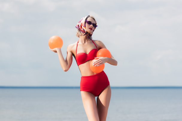 attractive elegant woman in red trendy bikini, sunglasses and silk scarf posing with balls at sea - Photo, image