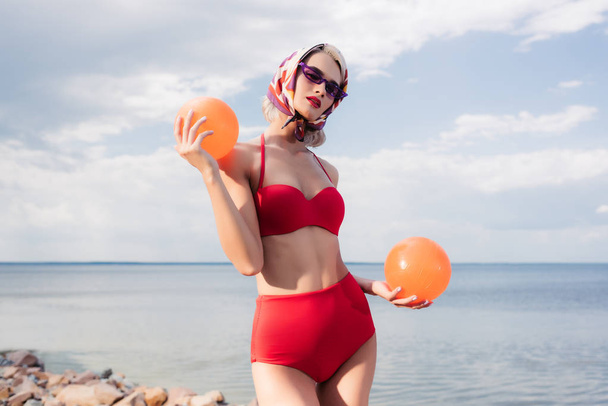 fashionable woman in red bikini, sunglasses and silk scarf posing with balls at sea - 写真・画像