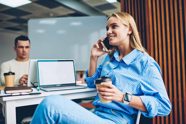 Cheerful blonde woman talking on mobile phone wit friend sitting at working place near laptop with mockup screen holding coffee cup,female employee having telephone conversation during work break - Zdjęcie, obraz