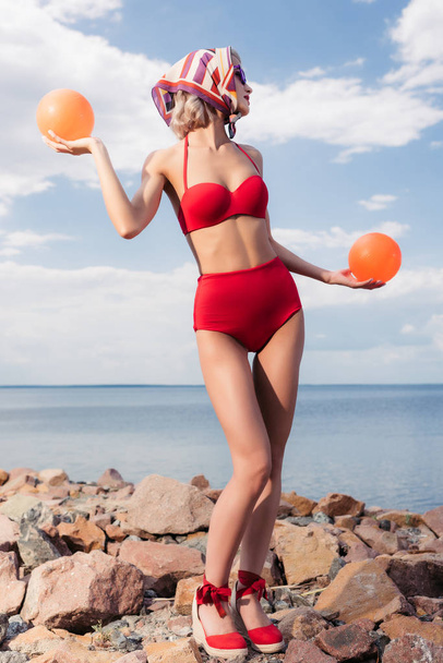 beautiful elegant girl in red bikini and silk scarf posing with balls on rocky shore - Photo, Image