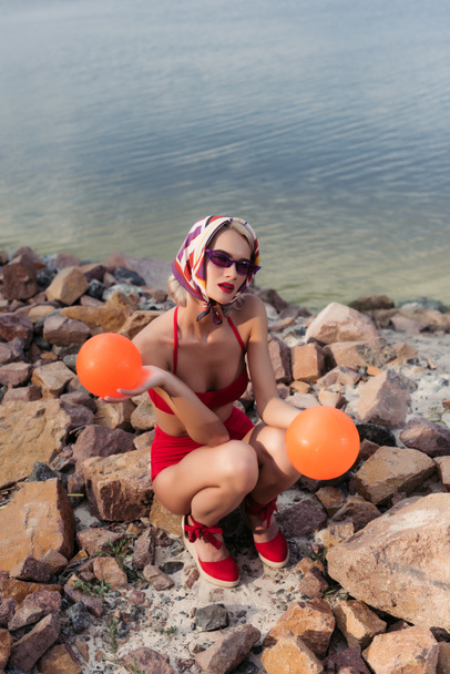 ελκυστική κοπέλα slim κόκκινο μαγιό, γυαλιά ηλίου και μεταξωτό μαντήλι ποζάρει με μπάλες στην βραχώδη παραλία - Φωτογραφία, εικόνα