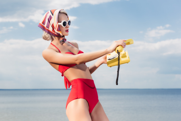 attraktive Frau im roten Bikini und Seidentuch posiert mit gelbem Telefon am Meer - Foto, Bild