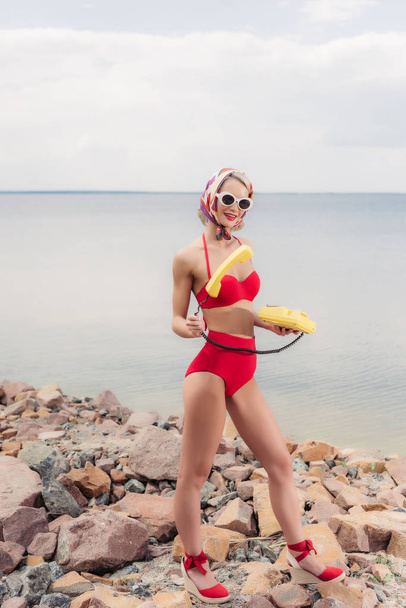 atractiva mujer en bikini vintage rojo posando con teléfono rotatorio amarillo en la playa rocosa cerca del mar
 - Foto, imagen