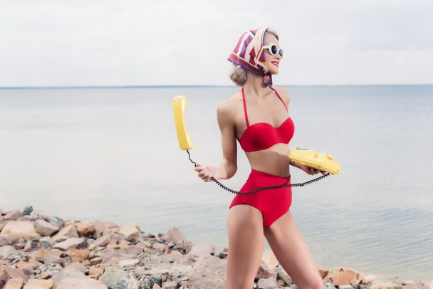 beautiful young woman in red bikini and silk scarf posing with yellow rotary telephone near the sea - Photo, Image