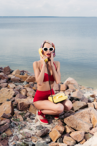 geschokt meisje in een rode bikini en zijden sjaal praten over vintage roterende telefoon - Foto, afbeelding