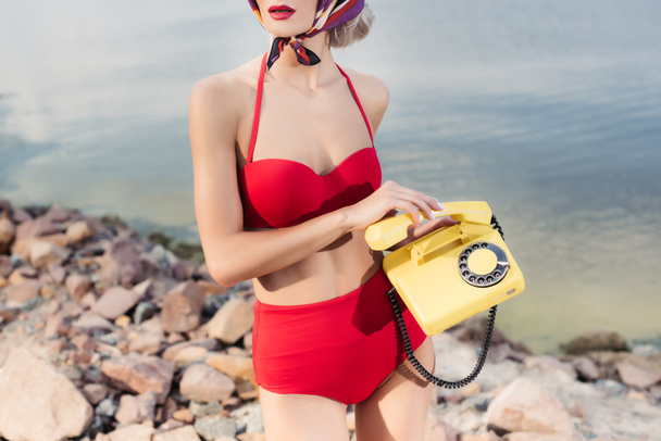 view modell vágott piros retro bikiniben pózol, rotációs telefon, sziklás strand - Fotó, kép