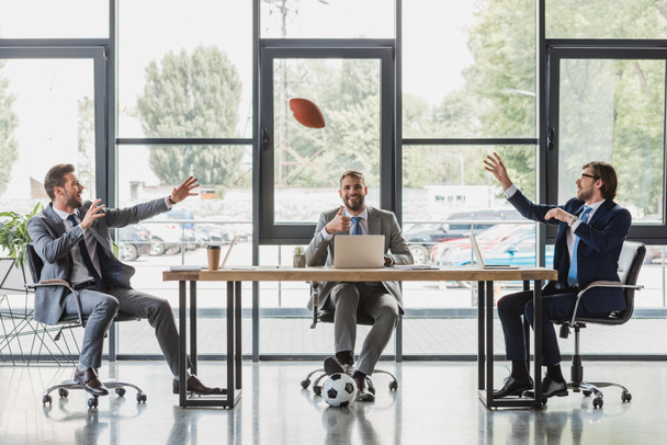 glimlachend jonge ondernemers met behulp van laptops en spelen met voetbal en rugby ballen in kantoor - Foto, afbeelding