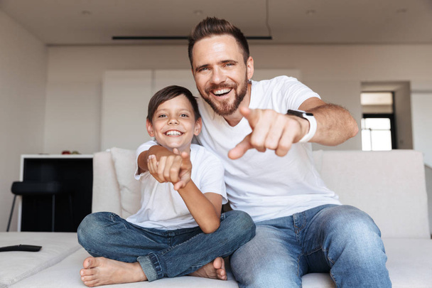 Image of cheerful glad man 30s and boy 8-10 laughing while pointing finger at you and sitting on sofa in apartment - Fotoğraf, Görsel