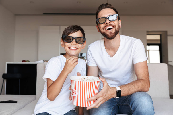 Portrait of joyful man 30s and boy 8-10 wearing 3d glasses eating popcorn and smiling while sitting on couch indoor and watching movie - Fotó, kép