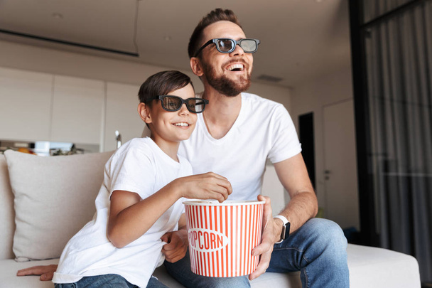 Image of caucasian man 30s and boy 8-10 wearing 3d glasses eating popcorn and smiling while sitting on couch at home and watching movie - Photo, image