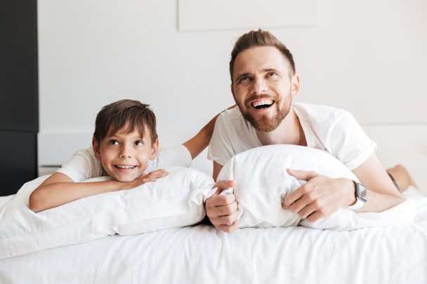 Изображение европейского мужчины 30 лет и мальчика 8-10 лет, смеющегося, лежа дома на кровати с белым бельем и глядя вверх
 - Фото, изображение