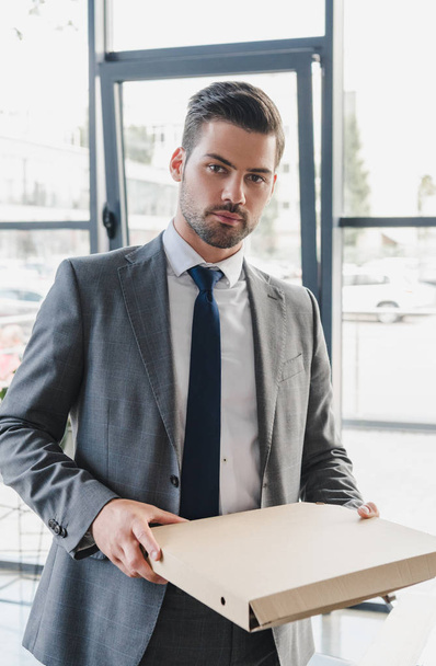handsome young businessman in suit holding pizza box and looking at camera in office - Φωτογραφία, εικόνα
