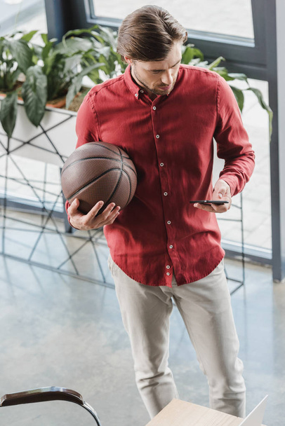 high angle view of businessman holding basketball ball and using smartphone in office - Foto, afbeelding