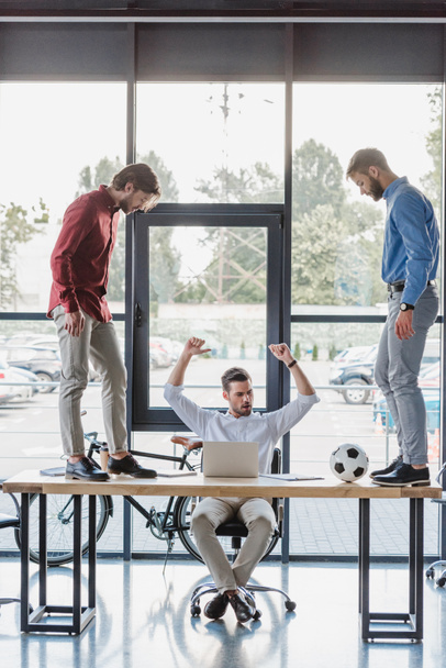 тріумфальний бізнесмен використовує ноутбук, коли колеги грають з футбольним м'ячем на столі
 - Фото, зображення