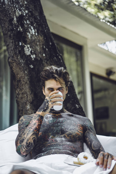 young handsome tattooed man having breakfast in bed in the open air outdoors in the garden - Foto, Imagen