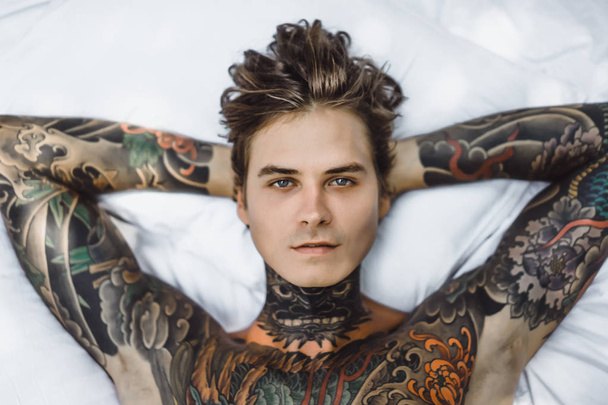 Handsome young tattooed man relaxing on white bed - Valokuva, kuva
