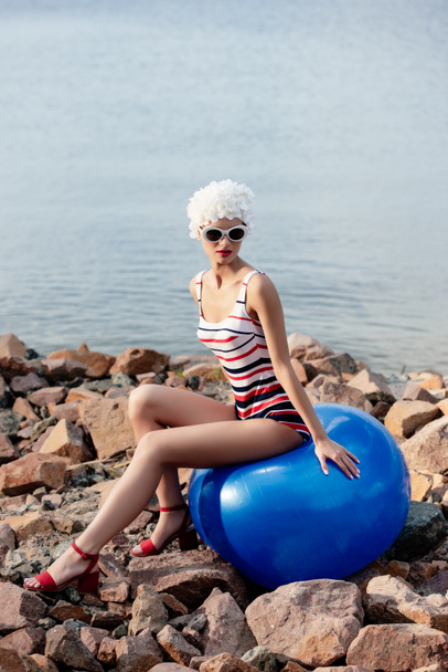 stylish attractive girl in vintage striped swimwear and sunglasses sitting on blue fit ball on rocky beach - Photo, Image