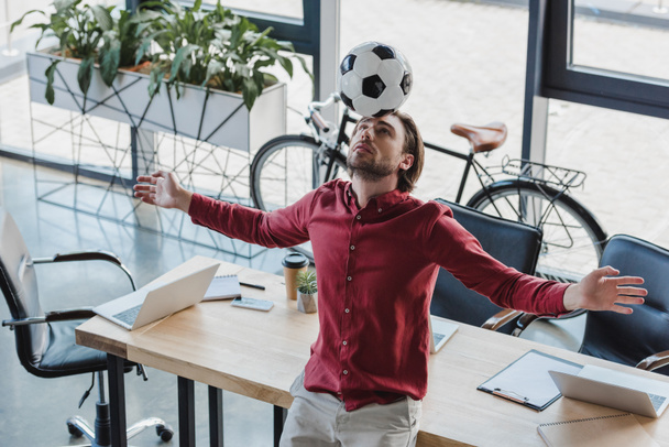 μεγάλη γωνία προβολής νεαρό επιχειρηματία εξισορρόπησης μπάλα ποδοσφαίρου στο κεφάλι στο γραφείο  - Φωτογραφία, εικόνα
