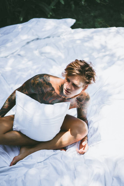 a young handsome tattooed man in the morning in bed tosses up the pillows. bed in the garden - Photo, image