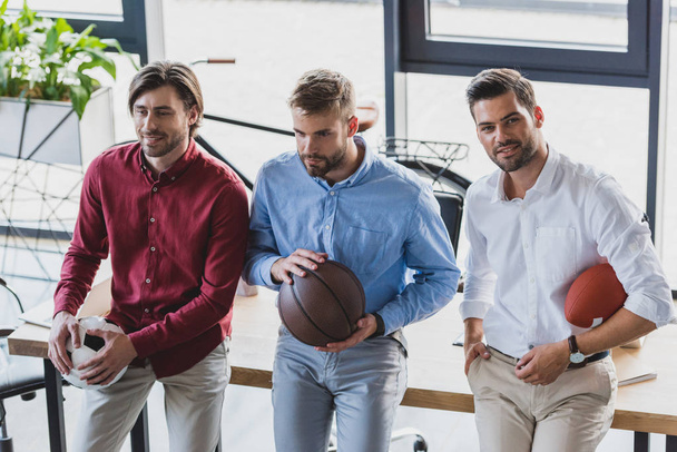 high angle view of young businessmen holding basketball, soccer and rugby balls in office - Photo, Image