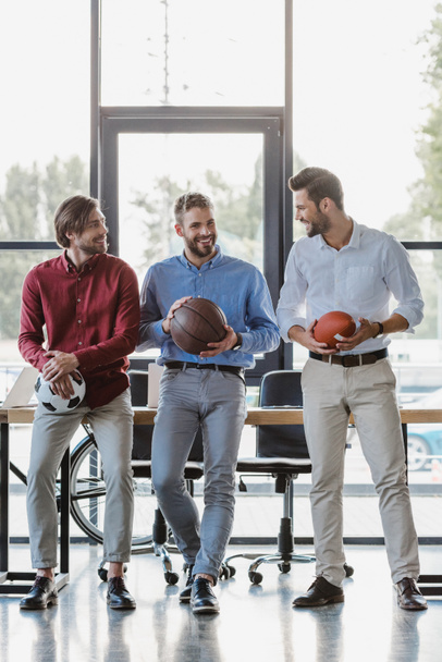 drei lächelnde junge Geschäftsleute mit Basketball-, Fußball- und Rugbybällen im Büro - Foto, Bild