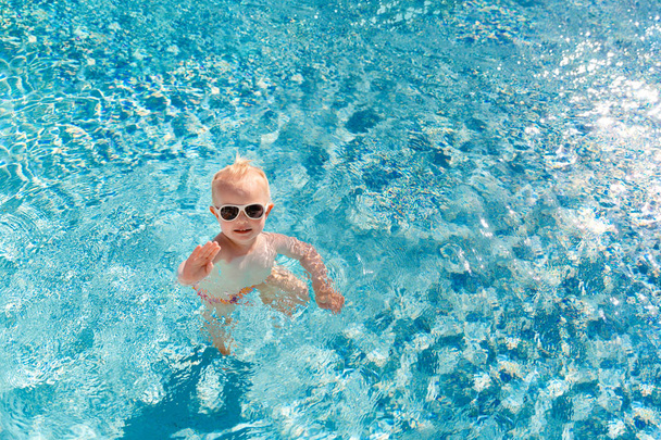Милый малыш в солнечных очках плавает в бассейне и машет рукой. Место для смс. Вид сверху
. - Фото, изображение