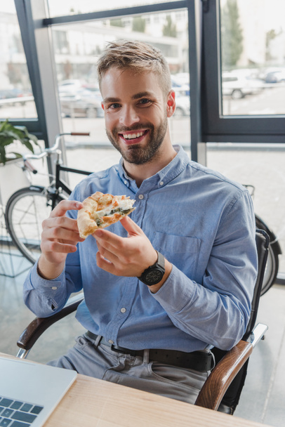 handsome young businessman holding pizza and smiling at camera in office - Foto, Imagem