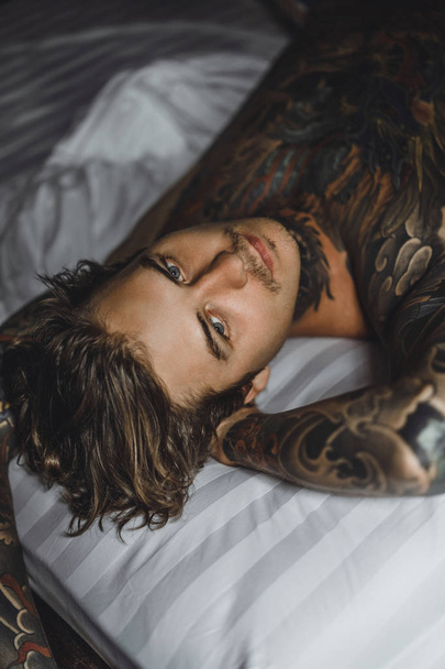 Handsome young tattooed man relaxing on white bed - Фото, изображение