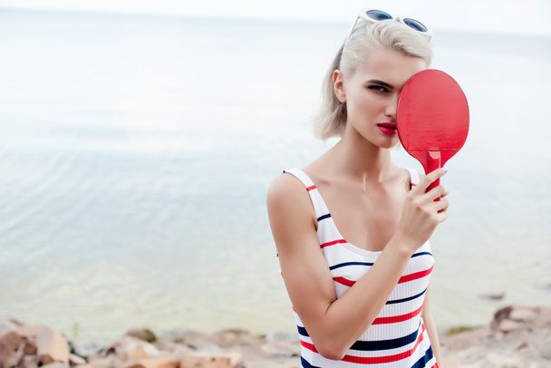 attractive sportswoman in retro striped swimsuit posing with ping pong racket on rocky beach - Foto, Bild