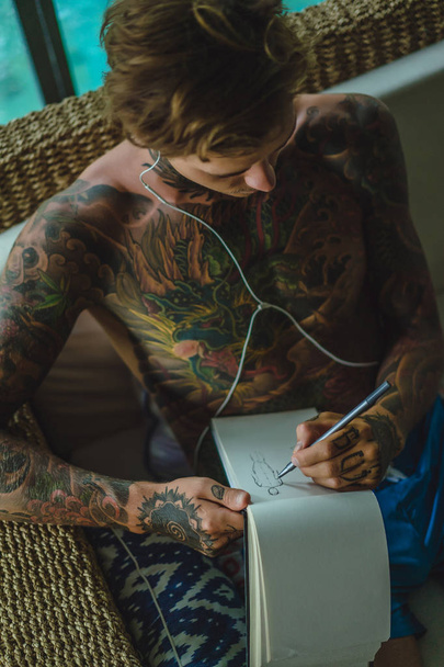 a young man in tattoos wearing headphones listens to music and draws a pencil in a notebook. Online training. - Zdjęcie, obraz