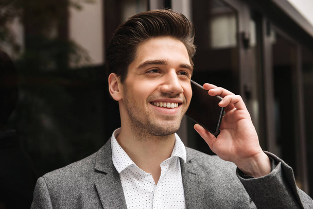 Close up of smiling young businessman talking on moible phone while standing at the glass doors outdoors - Fotoğraf, Görsel