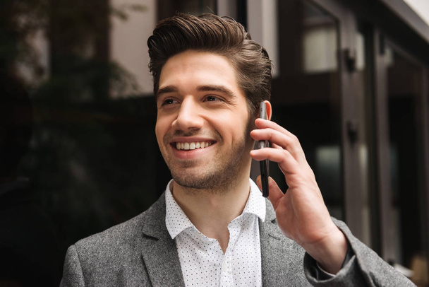 Close up of handsome young businessman talking on moible phone while standing at the glass doors outdoors - Фото, изображение