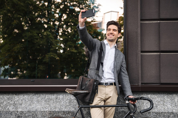 Усміхнений молодий бізнесмен, спираючись на стіну, стоїть на відкритому повітрі з велосипедом і приймає селфі
 - Фото, зображення