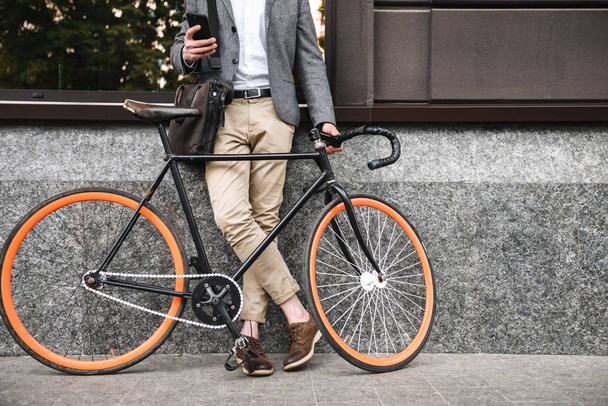 Cropped image of young businessman leaning on a wall while standing outdoors with a bicycle and using mobile phone - Foto, afbeelding