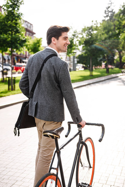 Achteraanzicht van lachende jonge zakenman wandelen met een fiets op de straat van de stad - Foto, afbeelding