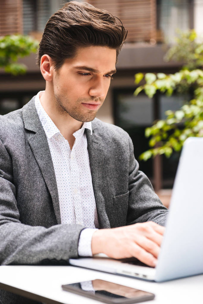 Focused young businessman dressed in suit working on laptop computer while sitting outdoors - 写真・画像