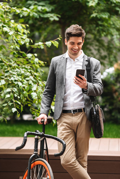 Attractive young businessman dressed in suit using mobile phone while standing outdoors with bicycle - Fotoğraf, Görsel