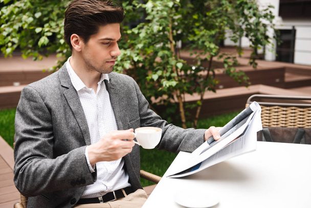 Confident young businessman dressed in suit drinking coffee while reading newspaper at the cafe outdoors - Photo, Image