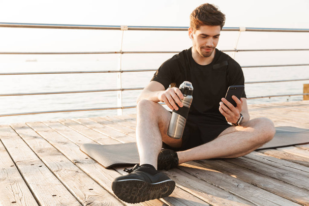 Foto de un joven deportista guapo sentado al aire libre en la playa escuchando música con auriculares usando un teléfono móvil sosteniendo la botella con agua
. - Foto, Imagen