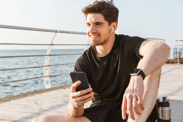 Photo of young happy sportsman outdoors on the beach listening music with earphones using mobile phone. - Foto, Bild