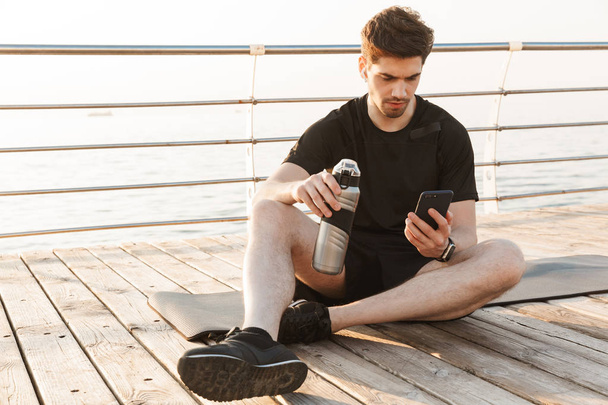 Photo of handsome young sportsman sitting outdoors on the beach listening music with earphones using mobile phone holding bottle with water. - Photo, Image