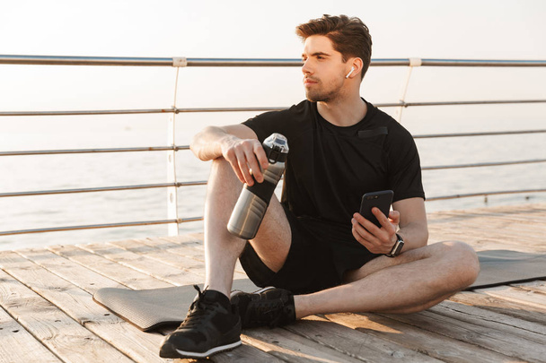 Photo of handsome young sportsman sitting outdoors on the beach listening music with earphones using mobile phone holding bottle with water. - Foto, Imagen