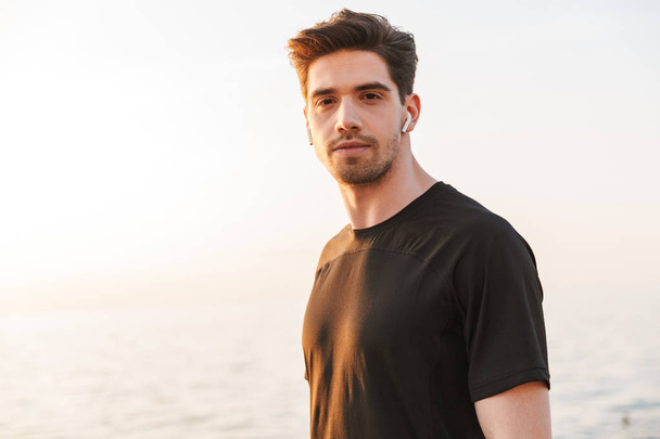 Confident young sportsman in black shirt in earphones standing at the seaside - Fotó, kép
