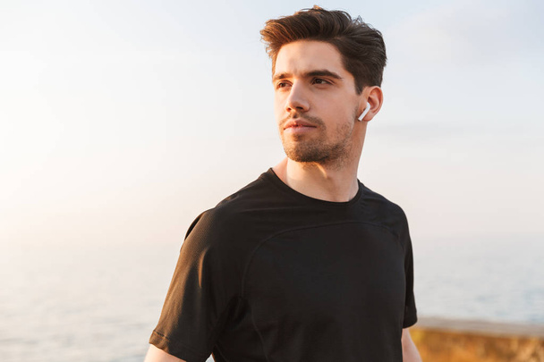 Motivated young sportsman in black shirt in earphones standing at the seaside - Fotoğraf, Görsel