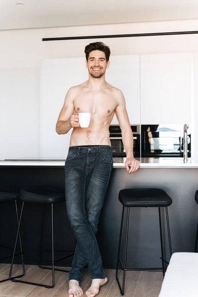 Image of handsome happy young man standing at the kitchen indoors drinking coffee looking camera. - 写真・画像