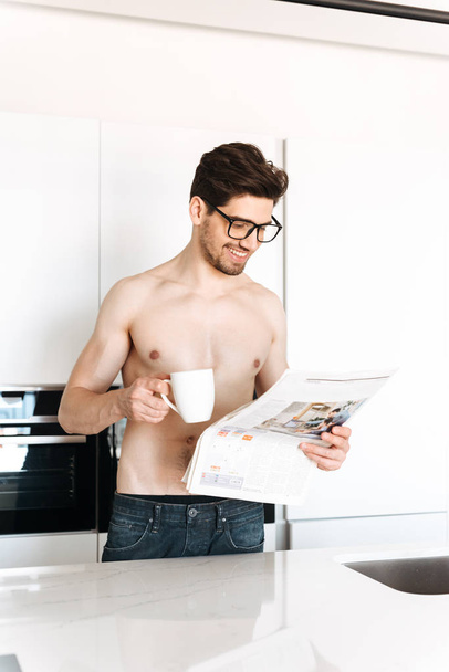 Image of young smiling man standing at the kitchen indoors reading newspaper drinking coffee looking aside. - Foto, immagini