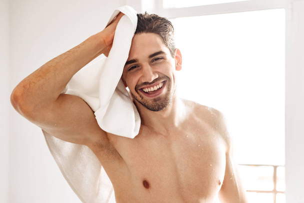 Foto de joven feliz hombre desnudo de pie en el interior en el baño con toalla. Mirando cámara
. - Foto, Imagen