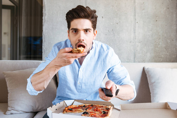 Shocked young man holding TV remote while sitting on a couch at home and eating pizza - Фото, зображення